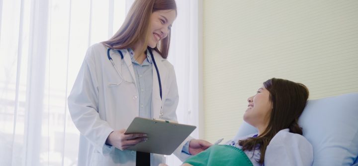 Beautiful smart Asian doctor and patient discussing and explaining something with clipboard in doctor hands while staying on Patient's bed at hospital. Medicine and health care concept.